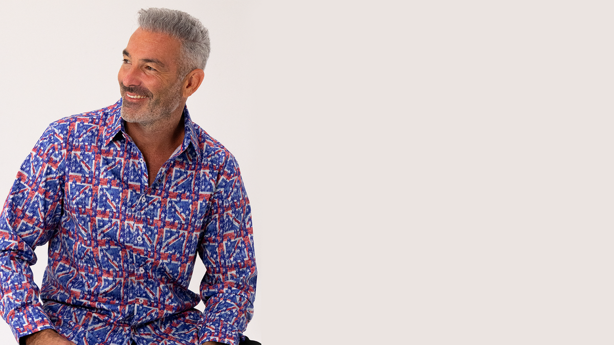 A smiling gray-haired man wearing a long-sleeve button-up shirt with a red, white, and blue geometric pattern, shown against a solid light beige background with negative space on the right.