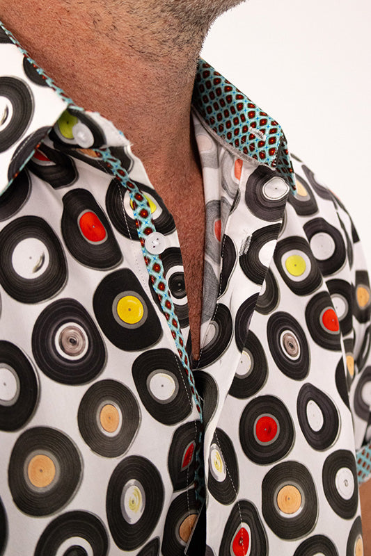 Close-up of a man wearing a white short-sleeve button-up shirt with a bold black vinyl record pattern and colorful record centers, featuring a patterned teal and black collar.