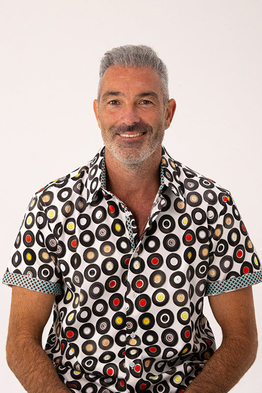 A smiling man with gray hair and a beard wearing a short-sleeve button-up shirt with a black, white, red, yellow, and beige circular vinyl record pattern, photographed against a plain white background.