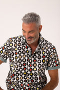 A man wearing a short-sleeve button-up shirt with a black, white circular vinyl record pattern, shown against a plain light background.