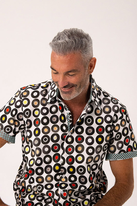 A man wearing a short-sleeve button-up shirt with a black, white circular vinyl record pattern, shown against a plain light background.