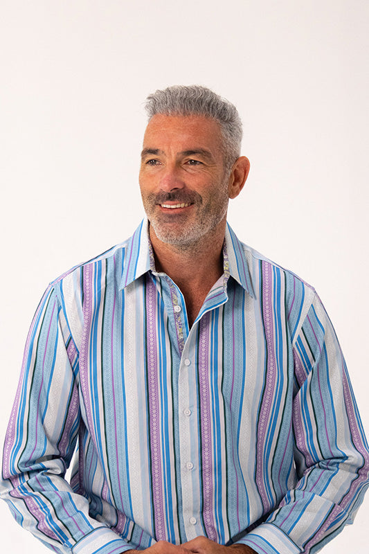 A smiling middle-aged man with gray hair wearing a long-sleeve button-up shirt with vertical stripes in blue, purple, and white, set against a plain white studio background.
