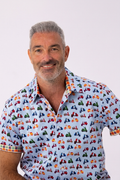 A smiling man with gray hair and a beard wearing a light blue short-sleeve button-up shirt with a colorful repeating scooter pattern, set against a plain light gray background.