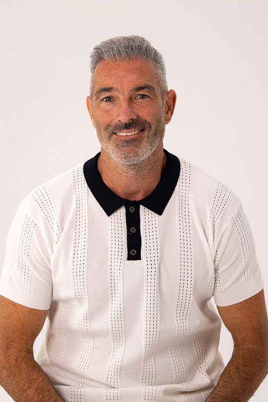 A smiling man with gray hair and a short beard wearing a white textured short-sleeve polo shirt with a black collar, photographed against a plain light background.