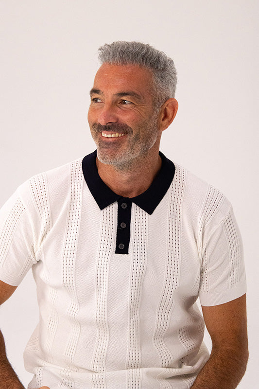 A smiling man with gray hair and a beard wearing a white short-sleeve knit polo shirt with a black collar and black buttons, photographed against a plain light background.
