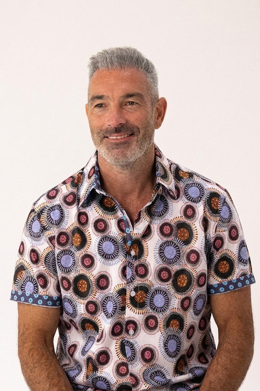 A smiling man with gray hair and a beard wearing a short-sleeve button-up shirt with a colorful circular geometric pattern, set against a plain light background.