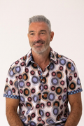 A smiling man with gray hair and a beard wearing a short-sleeve button-up shirt with a colorful circular geometric pattern, set against a plain light background.