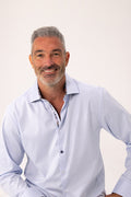A smiling man with gray hair and a short beard wearing a light blue long-sleeve button-down shirt, photographed against a plain off-white studio background.