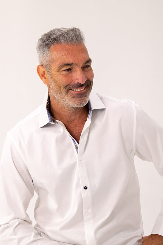 A smiling middle-aged man with gray hair and a short beard wearing a white long-sleeve button-down shirt, photographed against a plain light background.