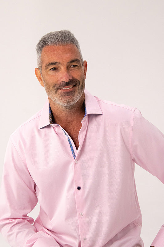 A smiling man with gray hair and a short beard wearing a light pink long-sleeve button-down shirt, photographed against a plain light background.