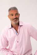 A smiling man with gray hair and a short beard wearing a light pink long-sleeve button-down shirt, photographed against a plain light background.