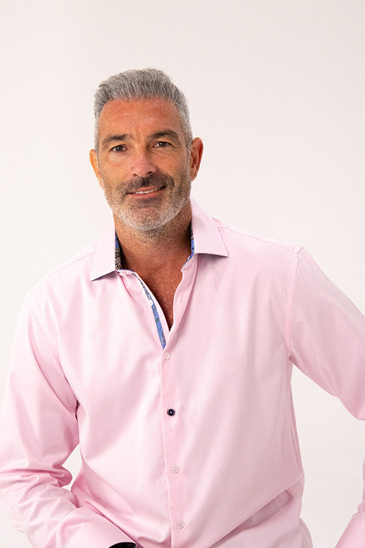 A man with gray hair and a short beard wearing a light pink long-sleeve button-down shirt, photographed against a plain light background.