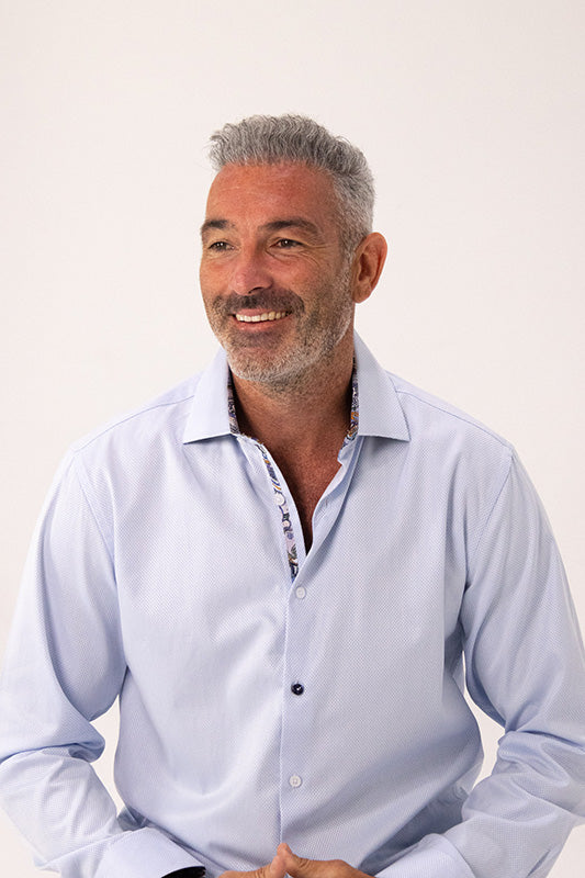 A smiling man with gray hair and a short beard wearing a light blue long-sleeve button-down shirt, photographed against a plain off-white background.