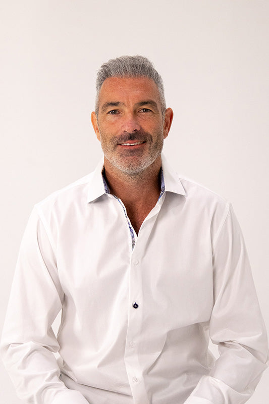 A smiling middle-aged man with gray hair and a graying beard, wearing a white long-sleeve button-down shirt with the top buttons undone, set against a plain light background.