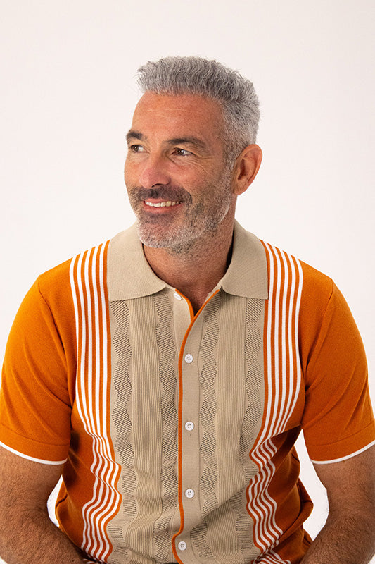 A smiling man with gray hair and a beard wearing a short-sleeve polo shirt with a beige textured front panel and orange sleeves, featuring orange and white vertical stripes on the shoulders and sides, against a plain white background.