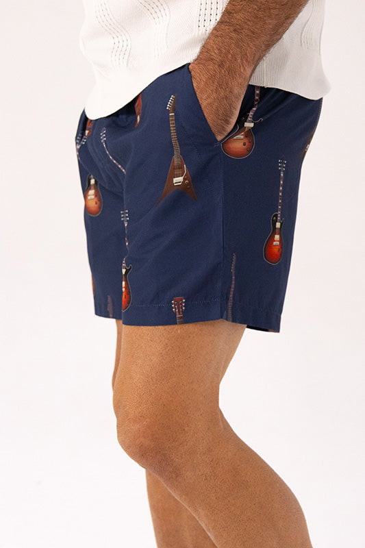A person wearing navy shorts with an allover print of guitars, paired with a white knit top, shown from the waist down against a plain white background.