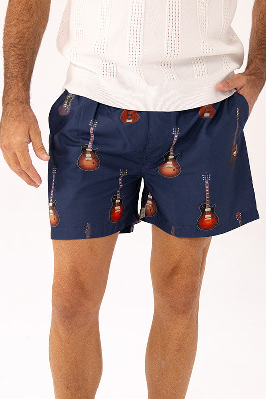 A man wearing navy blue swim short with a repeating guitar print, paired with a white knit top, shown from the chest down against a white studio background.