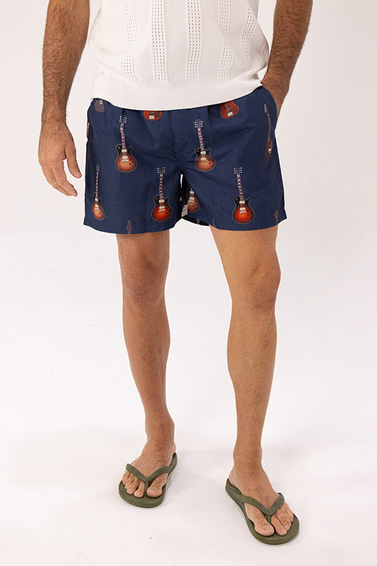 A man wearing navy blue shorts with a repeating guitar print, paired with a white knit top and olive green flip-flops, shown from the waist down against a white studio background.