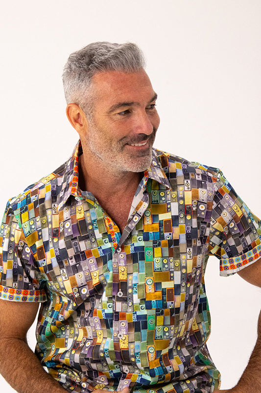 A smiling man with gray hair wearing a short-sleeve button-up shirt with a colorful geometric block pattern, photographed against a plain white background.