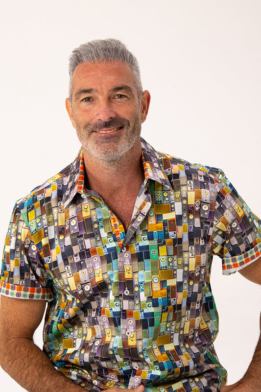 A smiling man with gray hair and a beard wearing a short-sleeve button-up shirt with a colorful geometric block pattern, photographed against a plain light background.