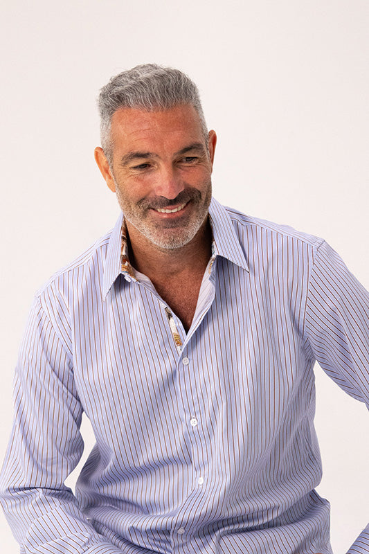 A smiling middle-aged man with gray hair and a short beard wearing a light blue and white vertically striped button-down shirt, photographed against a clean off-white studio background.