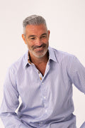 A smiling middle-aged man with gray hair and a short beard wearing a light blue and white vertically striped button-down shirt, photographed against a clean off-white studio background.