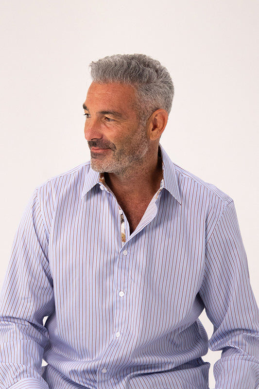 A man with gray hair and a beard wearing a light blue and white vertically striped button-down shirt, photographed against a plain light background.