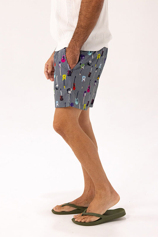 A man wearing gray pinstriped shorts with a colorful guitar print, a white knit top, and olive green flip-flops, shown from the waist down against a white studio background.