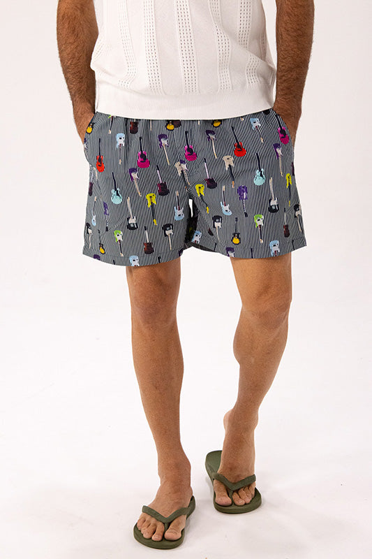 A man wearing gray shorts with a colorful guitar print, a white knit top, and green flip-flops, shown from the waist down against a white studio background.