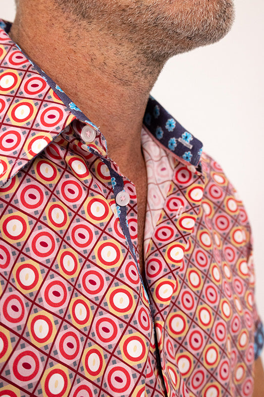 Close-up of a man wearing a short-sleeve button-up shirt with a colorful geometric pattern of red, orange, yellow, and white concentric circles on a pink background.