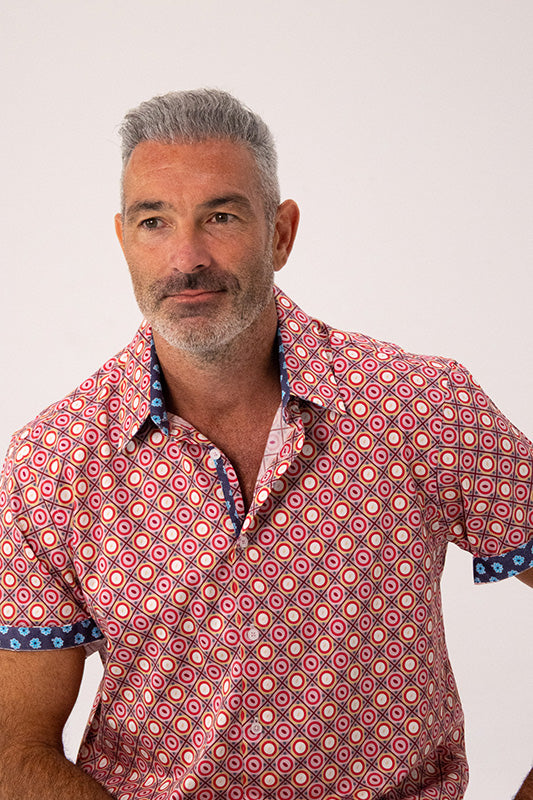 A man with grey hair wearing a short-sleeve button-up shirt with a red, pink, and white geometric pattern and blue patterned cuffs and collar trim, photographed against a plain light background.