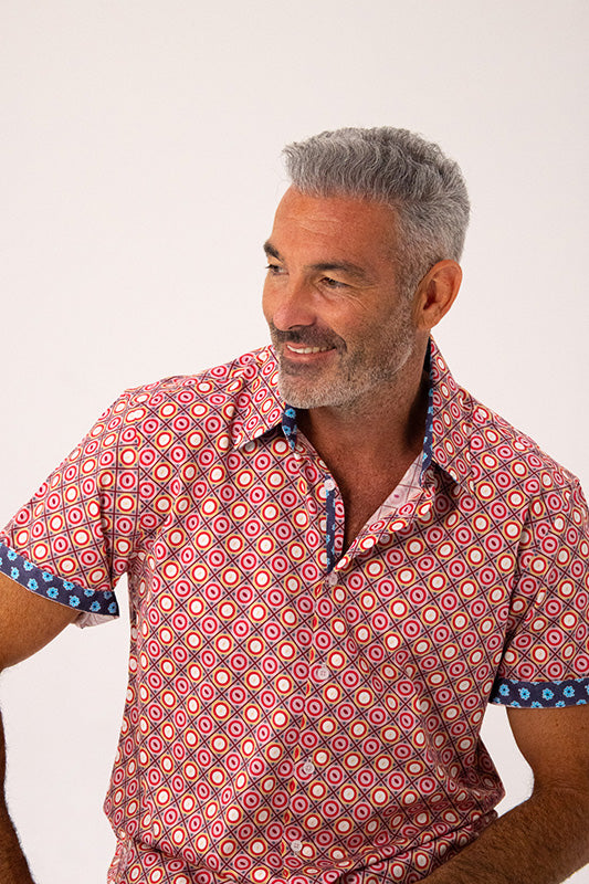 A man wearing a short-sleeve button-up shirt with a red, pink, and orange geometric pattern and blue patterned cuffs, photographed against a plain light background.