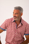 A man wearing a short-sleeve button-up shirt with a red, pink, and orange geometric pattern and blue patterned cuffs, photographed against a plain light background.