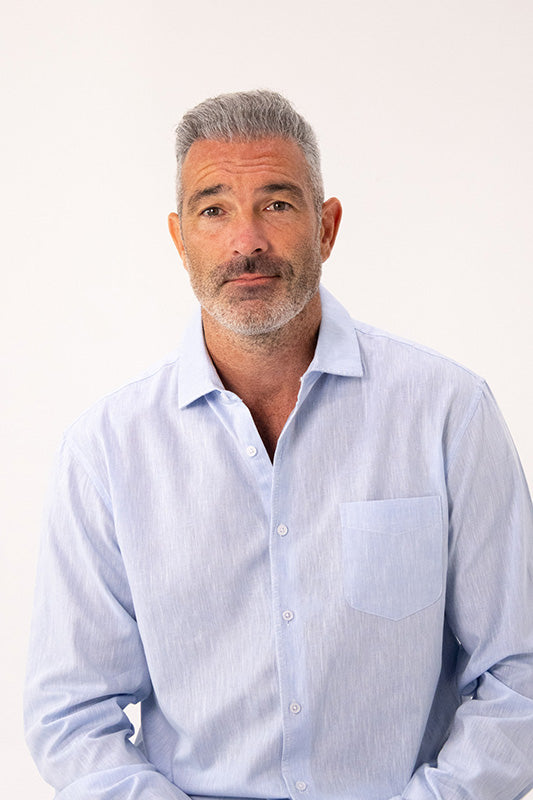 A man with gray hair and a short beard wearing a light blue long-sleeve button-down shirt, photographed against a plain white studio background.