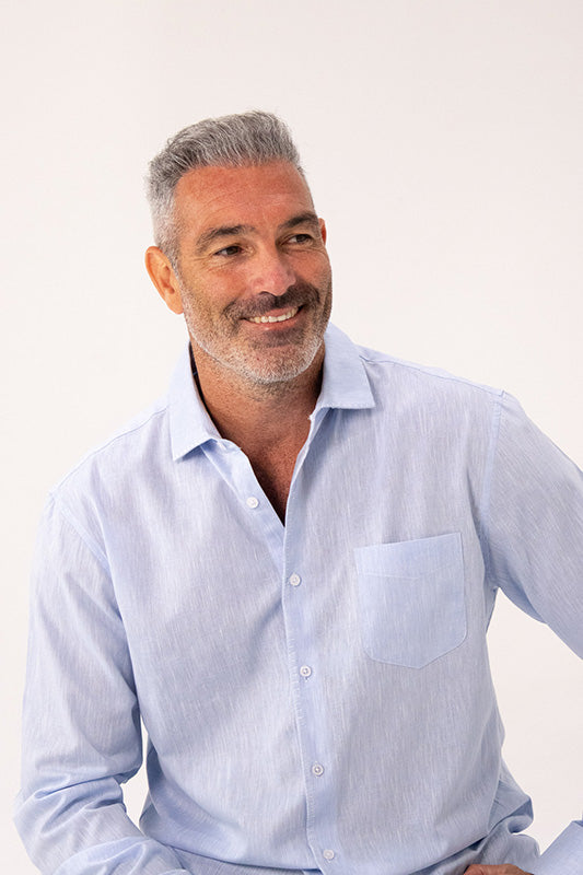 A smiling man with gray hair and a short beard wearing a light blue long-sleeve button-down shirt with a chest pocket, set against a plain off-white background.