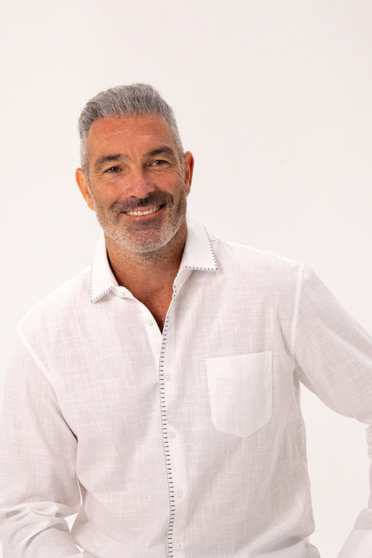 A smiling man with gray hair and a short beard wearing a white long-sleeve linen button-up shirt with decorative stitching, photographed against a clean white background.