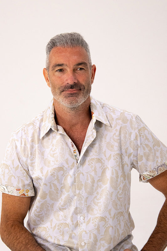 A man with gray hair and a beard wearing a short-sleeve button-up shirt with a beige and white paisley pattern, photographed against a plain light background.