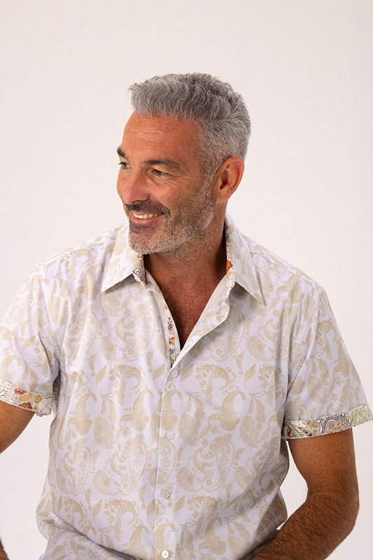 A smiling man with gray hair and a beard wearing a short-sleeve button-up shirt with a beige and white paisley pattern, photographed against a plain light background.