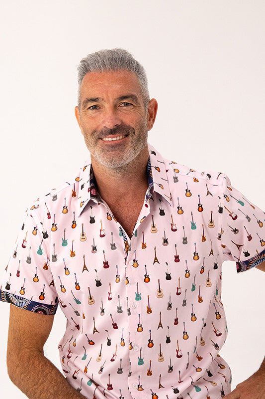 A smiling man with gray hair and a beard wearing a light pink short-sleeve button-up shirt covered in a colorful guitar print, set against a plain white background.