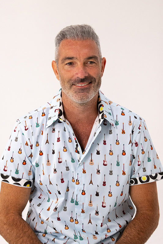 A smiling man with gray hair and a beard wearing a light blue short-sleeve button-up shirt with a repeating pattern of small guitars, photographed against a plain light background.