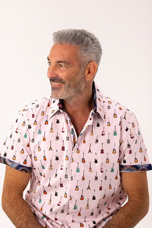 A man with gray hair and a beard wearing a light pink short-sleeve button-up shirt with a repeating pattern of small guitars in various colors, photographed against a plain white background.