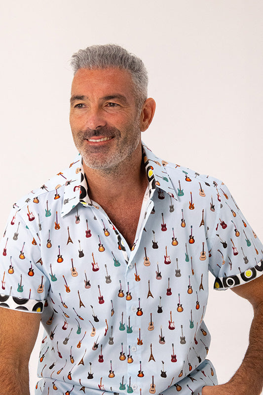 A smiling man with gray hair and a beard wearing a light blue short-sleeve button-up shirt covered in a colorful pattern of small musical instruments, photographed against a plain off-white background.