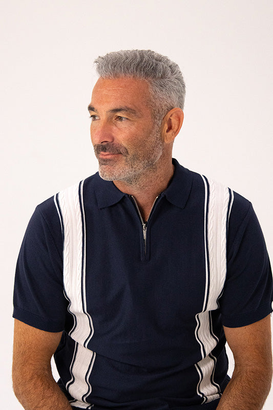 A man with gray hair and a beard wearing a navy blue polo shirt with white vertical textured stripes, photographed against a plain light background.