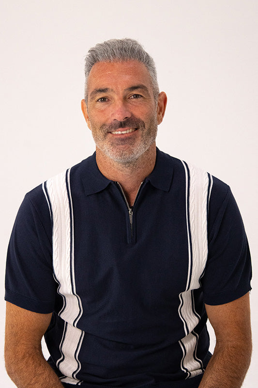 A smiling man with gray hair and a beard wearing a navy blue polo shirt with white vertical stripes, photographed against a plain light background.