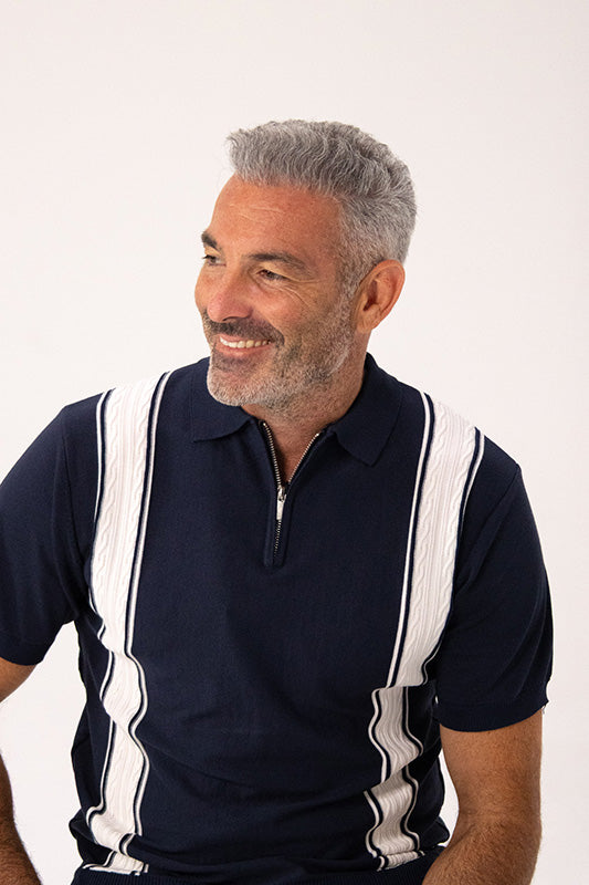A smiling man with gray hair and a short beard wearing a navy blue polo shirt with white vertical textured stripes on the shoulders and sides, photographed against a plain light background.