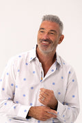 A smiling man with gray hair and a beard wearing a white long-sleeve button-up shirt with blue polka dots, photographed against a plain light background.