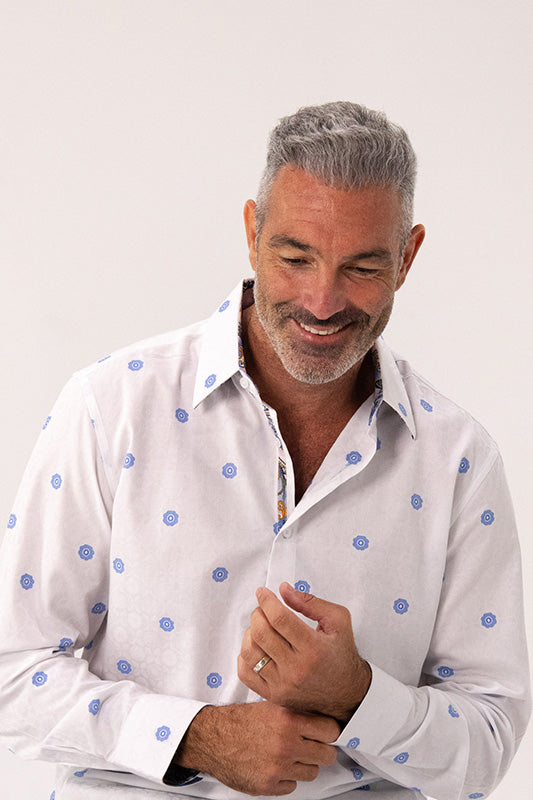 A smiling middle-aged man with gray hair and a beard, wearing a white long-sleeve button-down shirt with blue polka dots, adjusting his collar against a plain light background.